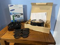 Four Samsung SSG-3570CR rechargeable 3D glasses placed on a wooden table with charging holders, cables, and original packaging.