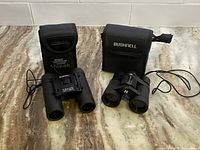Two pairs of Bushnell binoculars side by side with their carrying cases, showing the Powerview 12x25 and Powerview 4x30 models.
