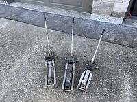 Three used hydraulic floor jacks placed upright on asphalt, showing long handles and metal bodies with visible signs of wear.
