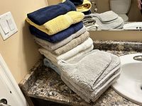 Stack of eight assorted towels folded and placed on a bathroom counter next to grey bathroom rugs.