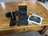 Two Amazon Fire TV Sticks with two remote controls and three H9 Mini wireless keyboards on wooden table, one keyboard in box, one in wrapper, and instruction manuals.