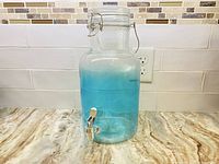 Front view of the blue tinted glass beverage dispenser showing the full container and silver spout at the bottom.