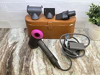 Main Dyson Supersonic hair dryer with attached power cord, leatherette case behind, and three styling attachments displayed on case.