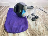 Massager lying on purple fabric pouch with four interchangeable massage heads displayed next to it on countertop.