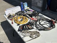 Full view of all electrical components, extension cords, battery cables, adaptors, automotive bulbs, and boxed aluminum flexible duct arranged on table outdoors.