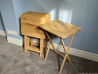 Five wooden folding snack tables stacked vertically and one opened to show structure and design. Photo shows natural light wood and foldable X-shaped legs.