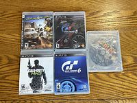 Front view of five PS3 game cases laid on wood surface featuring MotorStorm, Gran Turismo 5, Call of Duty MW3, Gran Turismo 6, with Need for Speed The Run disc out of case.