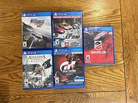 Top-down view of the five PS4 game cases arranged on floor surface showing cover art of Need for Speed Rivals, The Crew, Driveclub, Gran Turismo Sport and Assassin's Creed IV Black Flag.