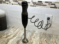 The KitchenAid immersion blender standing upright on a marble countertop with the power cord coiled behind it.