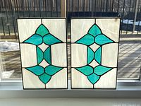 Two stained glass panels placed side by side on a white window sill with sunlight highlighting the turquoise and teal colors with white background.