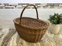 Front diagonal view of woven rattan basket with a single curved handle, placed on a marble countertop with tile backsplash.