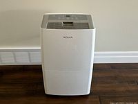 Front view of a white NOMA 50-pint portable dehumidifier sitting on a hardwood floor next to a wall vent, showing clean exterior and NOMA logo on the front.