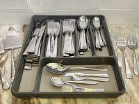 View of set of Oneida 90/15 stainless steel flatware pieces including forks, knives, spoons, pie servers arranged inside and outside a grey drawer organizer on a countertop.