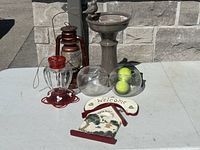 Full lot photo showing all five items on a white table outdoors in front of a stone wall: red metal lantern, glass hummingbird feeder, two glass bowls (one with tennis balls), gray bird bath with bird figurine, and wooden Welcome sign.