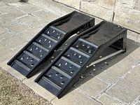 Pair of black vehicle ramps placed on a paved surface outdoors beside a stone wall, shown from front left angle highlighting textured surfaces with holes and raised side edges.