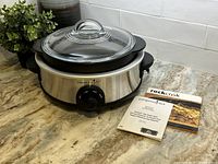 Slow cooker base with the Rockcrok pan on top and glass lid resting on the pan, with recipe booklet and manuals beside it.