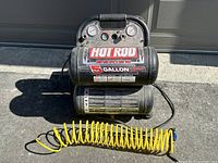 Front view of Powerbuilt Hot Rod Series 5-gallon dual-tank air compressor showing brand label, gauges, control knobs, and yellow coiled air hose in front.
