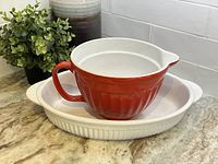 Red ceramic batter bowl with handle and spout sitting inside a white ribbed oval stoneware baking dish with handles on kitchen counter.