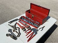 Red metal toolbox with various hand tools including sockets, ratchets, chain wrench, and pliers arranged on a table for display.