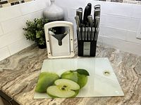Sabatier knife block with multiple knives and scissors, white bagel cutter, and rectangular glass apple cutting board on kitchen countertop.