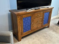 Solid oak Mission style buffet sideboard cabinet with blue maple leaf fabric panels on two doors and three central drawers, photographed with a TV on top in a room with beige carpet and light blue walls.