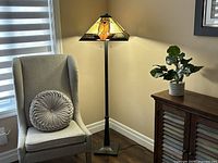 Full view showing the floor lamp next to a chair and wooden cabinet, lamp illuminated revealing stained glass shade colors and butterfly design.