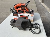 Three images showing two Black & Decker belt sanders, one with its original box and the other with a dust collection bag attached. Both tools have electrical cords and show signs of use.