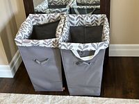 Front and top view of two grey fabric laundry hampers side by side, showing leaf print tops and front handles.
