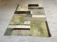 View of the full rectangular rug with geometric pattern in shades of green, brown, beige, and cream on grey floor.