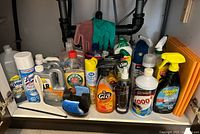 Photo showing a variety of used household cleaning products under a sink, including bottles of Dawn dish soap, Lysol spray, CLR cleaner, Murphy's Oil Soap, Orange Glo, Touch of Purple, 1000+ Stain Remover, Invisible Glass cleaner, Vim cleaner, along with cleaning gloves and sponge pads.