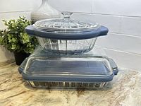 Stacked view of one round ribbed clear glass casserole dish with matching glass lid and blue plastic lid on top of one rectangular clear glass casserole dish with blue plastic lid.