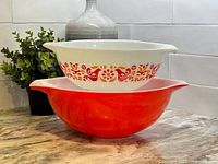 Two Pyrex mixing bowls stacked; larger solid red bowl on bottom and smaller white bowl with bird pattern on top.