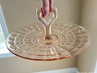 Clear view of the pink Depression glass tray held by the center handle, showing the radial ribbed pattern on the plate's surface and the geometric design of the handle.