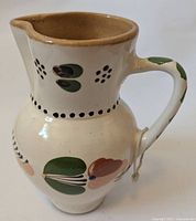 Front and side views of a ceramic jug showing traditional Hungarian folk art paintings with green, peach, and black floral patterns on a cream background.