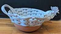 Front angled view showing the ceramic woven basket design, terracotta base, and rooster head with detailed features in glossy white ceramic.