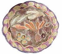 Top-down view of round ceramic art plate with raised vegetable sculptures and leaf border.