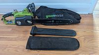 Greenworks electric chainsaw lying on floor with two black protective blade covers, one branded Greenworks.