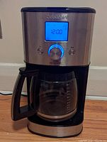 Front view of the coffee maker showing stainless steel finish, blue backlit LCD screen displaying time, glass carafe with handle and measurement markings.