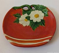 Front side of a red ceramic decorative plate with white daisies and green leaves and two holes for hanging.