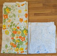 Folded view of both fabrics showing orange, yellow, green nasturtium design and blue/white floral design together on hardwood floor.