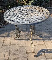 Photo showing round metal outdoor table with intricate floral openwork design on top, three legs with leaf details and surface rust.