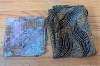 Two folded silk scarves, one with a pastel Prague themed pattern, the other with black and gold wavy lines, placed on wood floor.