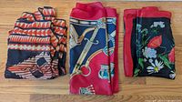 Three folded scarves and shawls arranged on wood floor showing different vibrant designs and fabrics