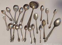 Photo of assortment of 20 used metal spoons and one serving fork in various sizes with diverse patterned handles