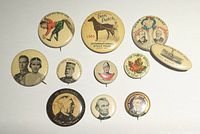 Top-down photo showing all 11 pins arranged on a white paper background, varying in size and displaying vintage promotional and political campaign imagery.