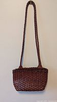 Full view of the brown woven leather shoulder bag hanging on a wall, showing the braided leather strap.
