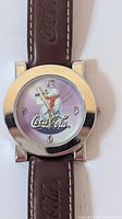 Front view of watch face showing bear graphic holding Coca-Cola and Coca-Cola logo, with silver bezel and brown embossed leather straps.