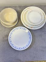 Photo showing three stacks of dishware: beige bowls and plates, white plates with orange and gray floral rim, and white plate with blue floral rim in front.