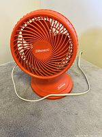 Orange Sunbeam electric fan showing front grille and base on a gray surface with attached power cord.