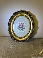 Framed cross stitch with pink rose and green leaves in an ornate oval frame, placed on a grey surface, side view.